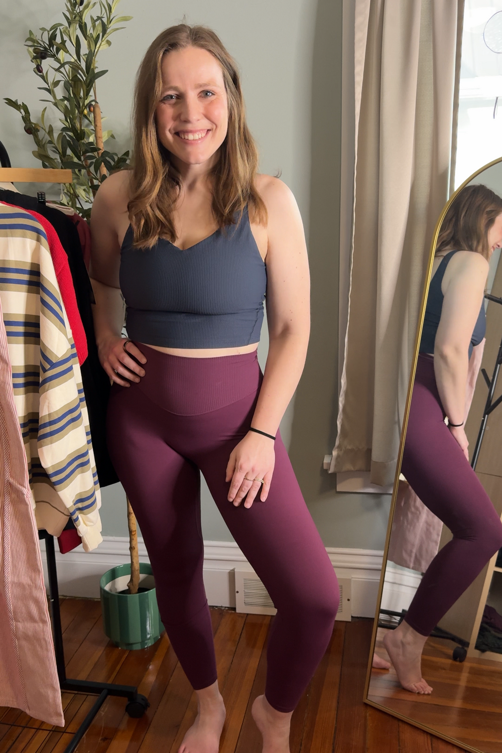 Woman wearing a dark gray sports bra and purple leggings standing in a room with a mirror and clothing rack.