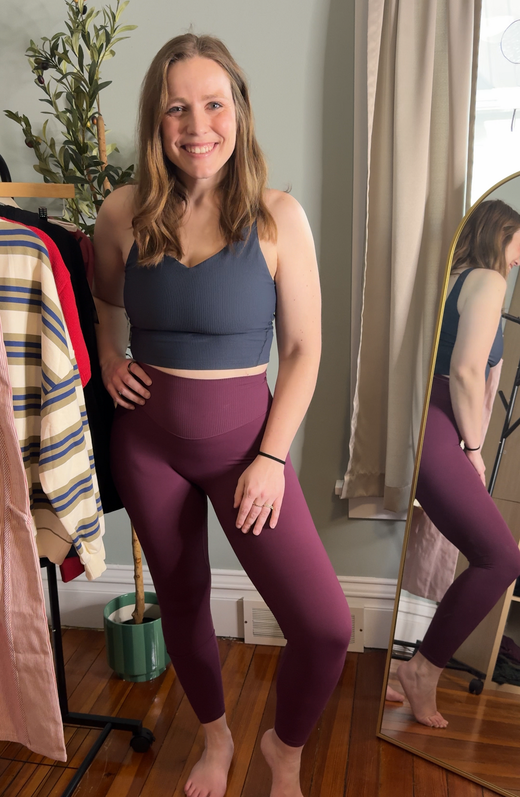 Woman wearing a dark gray sports bra and purple leggings standing in a room with a mirror and clothing rack.